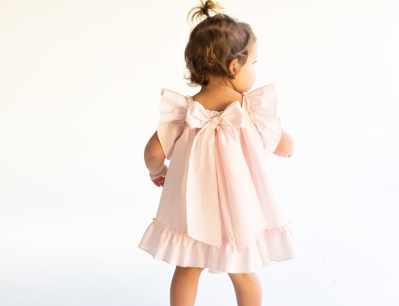 May include: A little girl wearing a pink dress with a large bow on the back. The dress has ruffled sleeves and a ruffled hem.