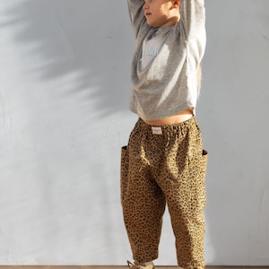 May include: A child wearing a gray long-sleeved shirt with a light blue graphic and leopard print pants. The pants have large pockets. The child is also wearing beige and white sneakers.