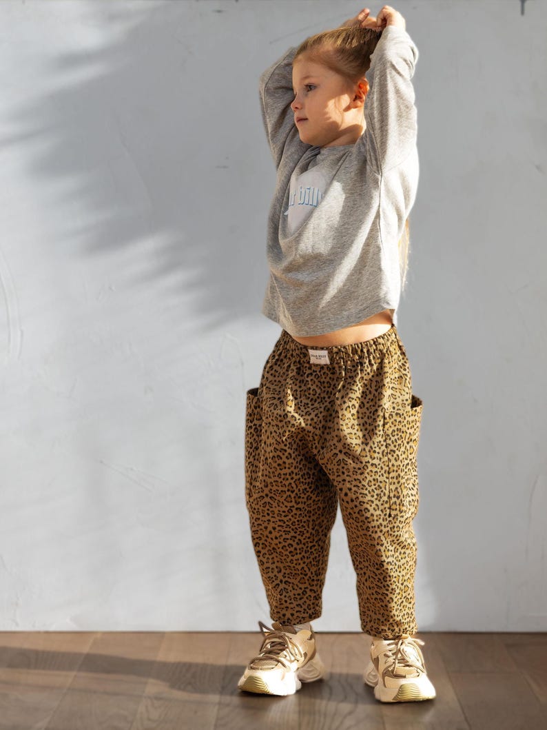 May include: A child wearing a gray long-sleeved shirt with a light blue graphic and leopard print pants. The pants have large pockets. The child is also wearing beige and white sneakers.