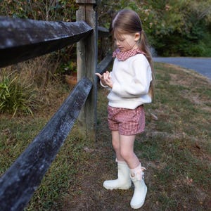 May include: A young person wearing a white sweater with a red and white checkered shorts. The outfit is paired with white rain boots. The person is standing next to a wooden fence in a grassy area.