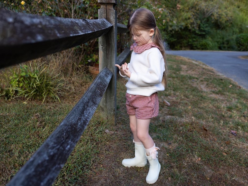 May include: A young person wearing a white sweater with a red and white checkered shorts. The outfit is paired with white rain boots. The person is standing next to a wooden fence in a grassy area.