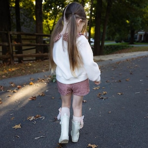 May include: A young person wearing a white sweater with a patterned collar and red and white checkered shorts. They are wearing white boots and walking on a paved road. The background includes trees and a wooden fence.
