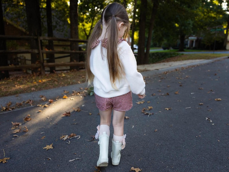 May include: A young person wearing a white sweater with a patterned collar and red and white checkered shorts. They are wearing white boots and walking on a paved road. The background includes trees and a wooden fence.