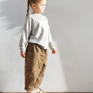 May include: A young child wearing a gray long-sleeved sweatshirt and leopard print pants. The child is also wearing white and tan sneakers. The outfit is casual and suitable for everyday wear.