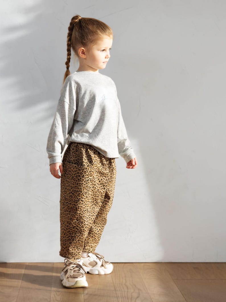 May include: A young child wearing a gray long-sleeved sweatshirt and leopard print pants. The child is also wearing white and tan sneakers. The outfit is casual and suitable for everyday wear.