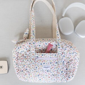 May include: A white floral print duffel bag with a zippered front pocket and cream-colored handles. The bag has a quilted pattern and contains a pink tube of lip balm. A pair of white headphones and a compact are also visible.