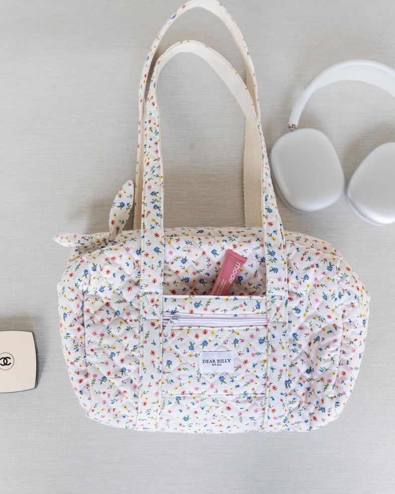 May include: A white floral print duffel bag with a zippered front pocket and cream-colored handles. The bag has a quilted pattern and contains a pink tube of lip balm. A pair of white headphones and a compact are also visible.