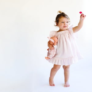 May include: A young girl in a pink dress holds a doll and a pink flower. She is standing on a white background.