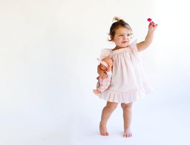 May include: A young girl in a pink dress holds a doll and a pink flower. She is standing on a white background.