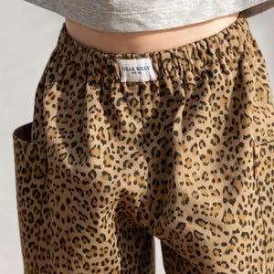 May include: Close-up of tan pants with a leopard print pattern. The pants have a drawstring waist and pockets. A small white tag with the text "DEAR BILLY" is visible on the waistband. The pants are paired with a gray top.