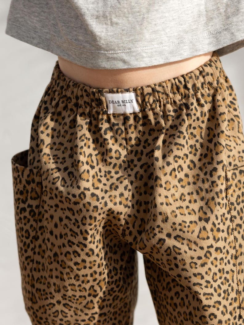 May include: Close-up of tan pants with a leopard print pattern. The pants have a drawstring waist and pockets. A small white tag with the text "DEAR BILLY" is visible on the waistband. The pants are paired with a gray top.