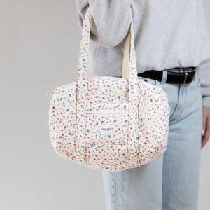 May include: A quilted floral print handbag with a white background and colorful flowers. The bag has a zippered front pocket and two handles. The text "DEAR BILLY" is on the front of the bag.