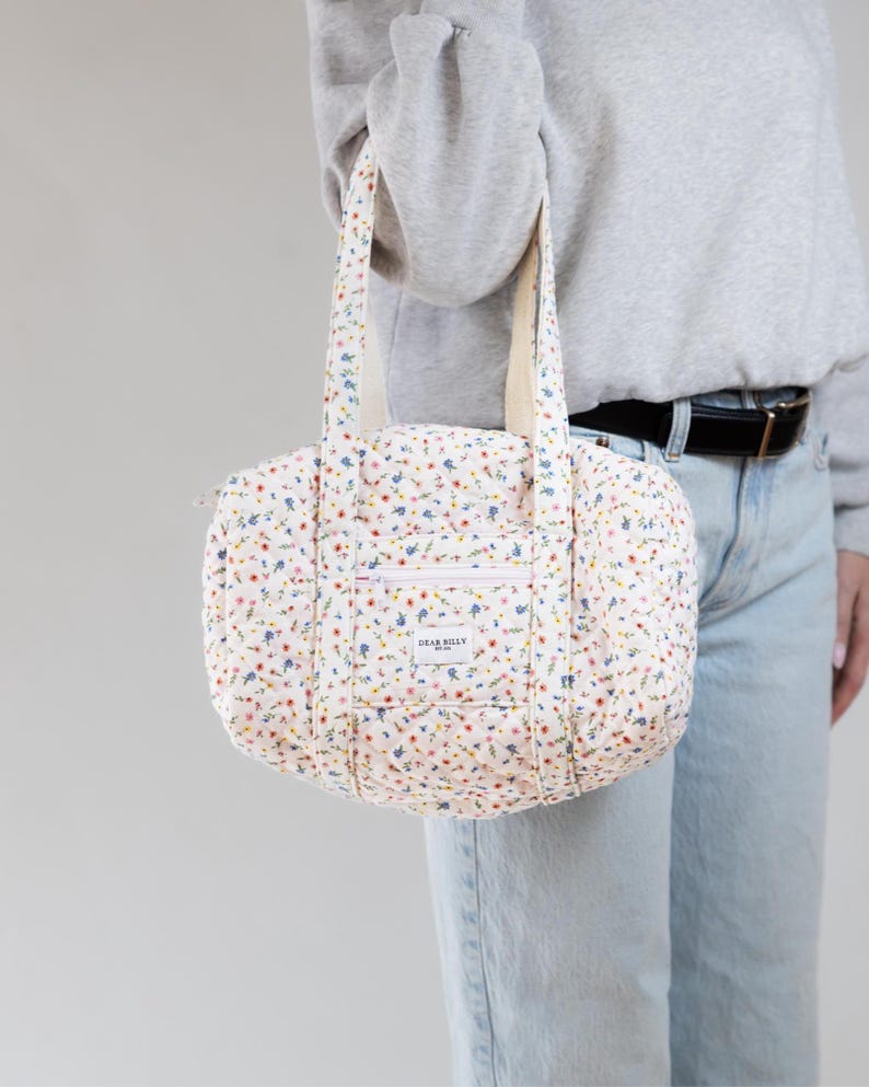 May include: A quilted floral print handbag with a white background and colorful flowers. The bag has a zippered front pocket and two handles. The text "DEAR BILLY" is on the front of the bag.