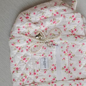 May include: A quilted white fabric item with a pink floral pattern and a white zipper. A pearl necklace with a heart-shaped clasp and pendant is draped across the item. The label reads "DEAR BILLY EST. 2018".