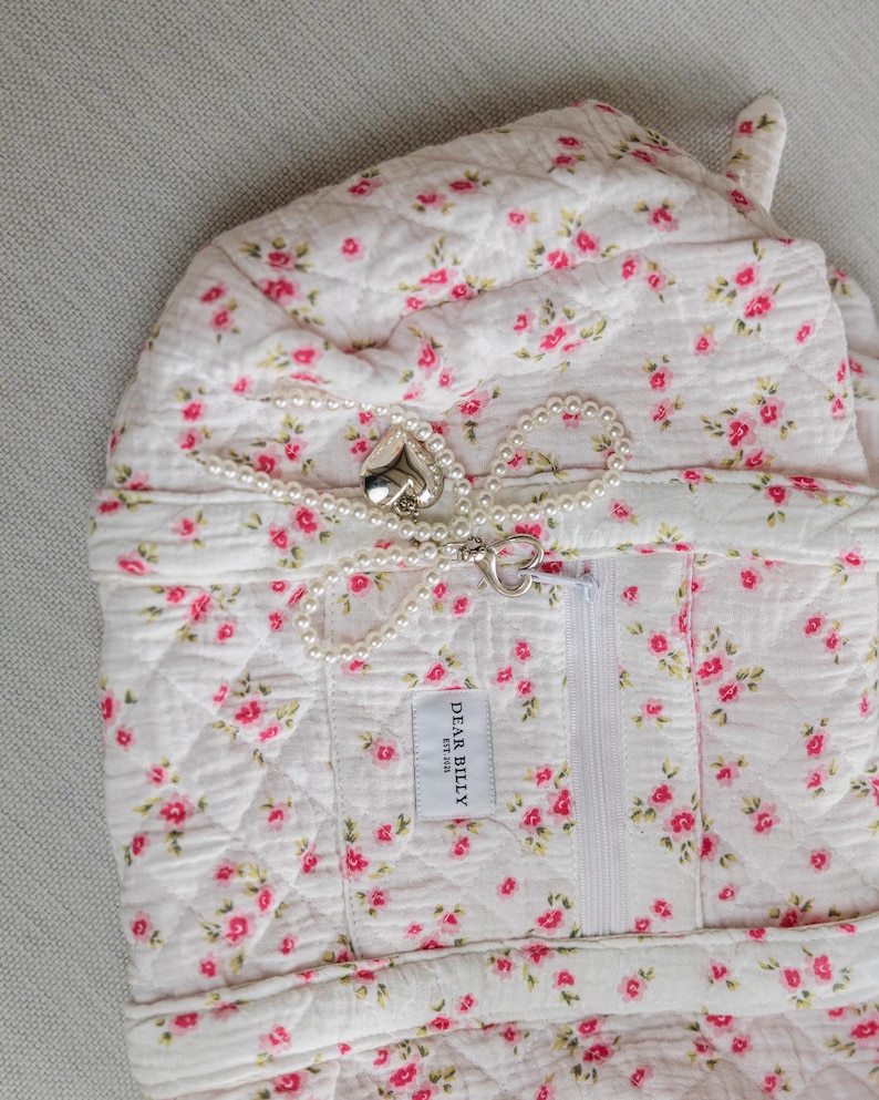 May include: A quilted white fabric item with a pink floral pattern and a white zipper. A pearl necklace with a heart-shaped clasp and pendant is draped across the item. The label reads "DEAR BILLY EST. 2018".