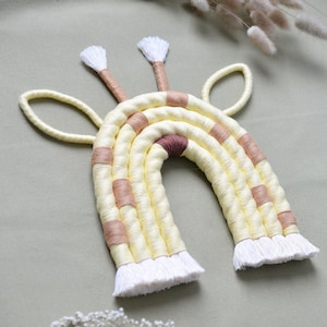 Puede incluir: Un tapiz de macramé arcoíris amarillo y marrón con borlas blancas. El arcoíris está hecho de hilo tejido y tiene una forma de jirafa con dos orejas.