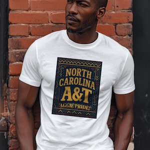 Könnte beinhalten: Weißes T-Shirt mit einem rechteckigen Design, das "NORTH CAROLINA A&T AGGIE PRIDE" in Gold und Marineblau zeigt. Das Design wird von einem dekorativen Rand mit geometrischen Mustern umrahmt. Das T-Shirt hat kurze Ärmel.