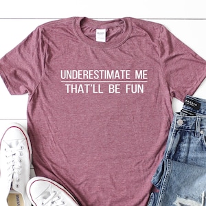 May include: Heather maroon t-shirt with the text "UNDERESTIMATE ME THAT'LL BE FUN" in white. The shirt is paired with blue jeans and white sneakers.