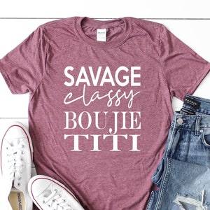 May include: Heather maroon t-shirt with white text that reads "Savage classy boujie titti". The shirt is shown on a white surface with a pair of white sneakers and blue jeans.