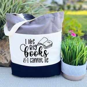 May include: A white canvas tote bag with black handles and a black bottom. The bag has a gray stripe across the middle and a black graphic that says "I like big books & I cannot lie".