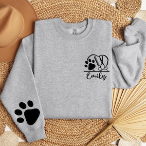 May include: Gray sweatshirt with a black paw print and scissors design on the front. The text "Emily" is below the design. The sweatshirt has a black paw print on the left sleeve.