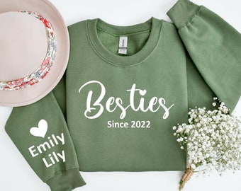 May include: A green sweatshirt with the text "Besties Since 2022" and a small heart on the front. The left sleeve has a heart and the text "Emily Lily" on it.
