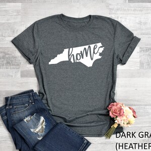 K&ouml;nnte beinhalten: Dunkelgraues T-Shirt mit einem wei&szlig;en Umriss des Bundesstaates North Carolina und dem Wort "home" innerhalb des Bundesstaates.