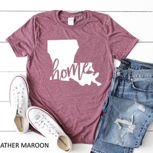 Pu&ograve; includere: T-shirt color tortora con un contorno bianco dello stato della Louisiana e la parola "home" scritta in corsivo.