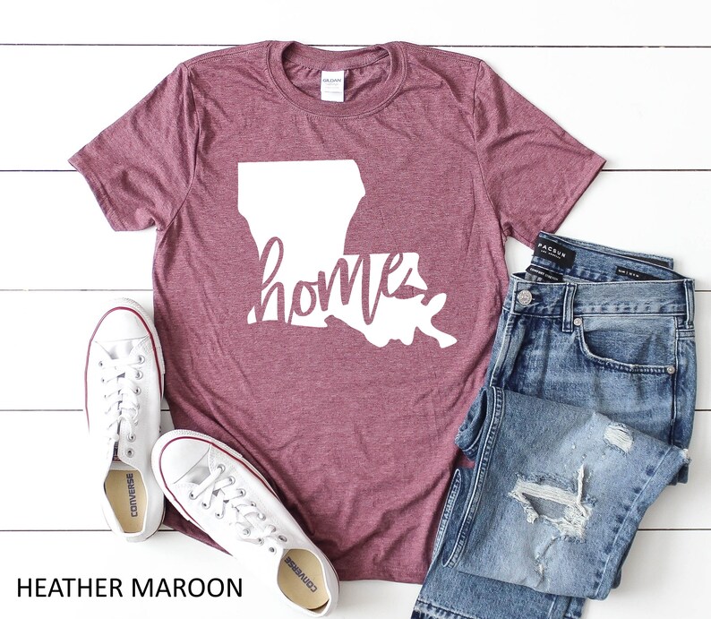 Pu&ograve; includere: T-shirt color tortora con un contorno bianco dello stato della Louisiana e la parola "home" scritta in corsivo.