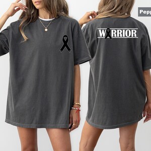 May include: Pepper-colored t-shirt with a black ribbon graphic on the front and back. The back of the shirt features the word "WARRIOR" in white block letters above the ribbon. The shirt has a relaxed fit.