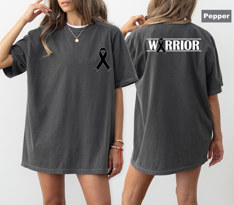 May include: Pepper-colored t-shirt with a black ribbon graphic on the front and back. The back of the shirt features the word "WARRIOR" in white block letters above the ribbon. The shirt has a relaxed fit.