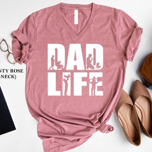 Peut inclure: T-shirt col en V rose poussi&eacute;reux avec le texte "DAD LIFE" en blanc. Le texte est entour&eacute; de silhouettes d'un p&egrave;re interagissant avec ses enfants.