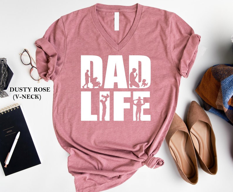 Peut inclure: T-shirt col en V rose poussi&eacute;reux avec le texte "DAD LIFE" en blanc. Le texte est entour&eacute; de silhouettes d'un p&egrave;re interagissant avec ses enfants.