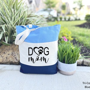 May include: A blue, white and navy blue canvas tote bag with the text "DOG mom" in black lettering. The "O" in "DOG" is replaced with a heart with a paw print inside.