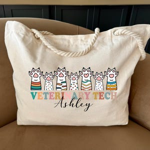 May include: Cream-colored tote bag with a rope handle. The bag features a colorful paw print design with the words "Veterinary Tech" and a name in a script font. The bag is sitting on a tan couch.