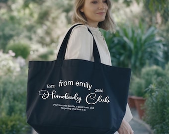 May include: A black tote bag with white text. The text reads "EST. from emily 2026 Homebody Club" and "your favourite candle, a good book, and forgetting what time it is". The bag has two black handles.