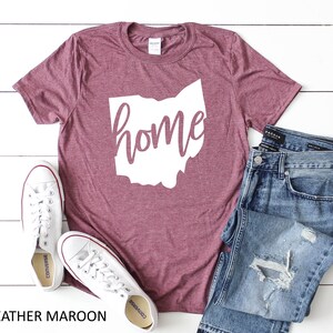 K&ouml;nnte beinhalten: Heather-marronfarbenes T-Shirt mit einer wei&szlig;en Umrandung des Bundesstaates Ohio und dem Wort "home" darin. Das Shirt liegt auf einer wei&szlig;en Holzoberfl&auml;che mit einem Paar wei&szlig;en Converse-Turnschuhen und einer blauen Jeans.