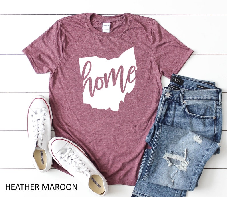 K&ouml;nnte beinhalten: Heather-marronfarbenes T-Shirt mit einer wei&szlig;en Umrandung des Bundesstaates Ohio und dem Wort "home" darin. Das Shirt liegt auf einer wei&szlig;en Holzoberfl&auml;che mit einem Paar wei&szlig;en Converse-Turnschuhen und einer blauen Jeans.