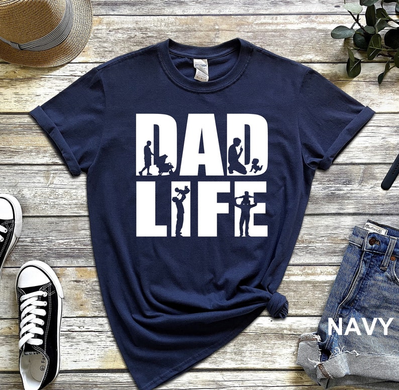 Peut inclure: T-shirt bleu marine avec un texte blanc qui dit "DAD LIFE". Le texte est compos&eacute; de silhouettes d'un p&egrave;re &agrave; diff&eacute;rents stades de la paternit&eacute;, de la tenue d'un b&eacute;b&eacute; &agrave; la  jouer avec un enfant.