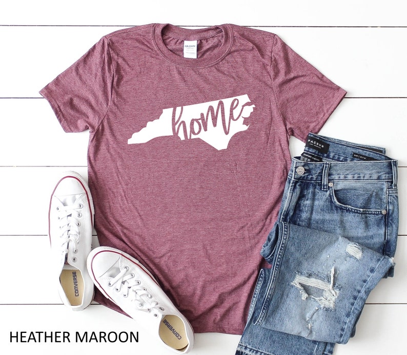 K&ouml;nnte beinhalten: Heather-marronfarbenes T-Shirt mit einem wei&szlig;en Umriss des Bundesstaates North Carolina und dem Wort "home" innerhalb des Bundesstaates. Das Shirt liegt auf einer wei&szlig;en Holzoberfl&auml;che mit einem Paar wei&szlig;en Converse-Schuhen und einer blauen Jeans mit Rissen.