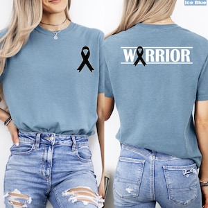 May include: Ice blue t-shirt with a black ribbon graphic on the front and back. The back of the shirt features the word "WARRIOR" in white block letters above the ribbon. The shirt is made of a soft material.