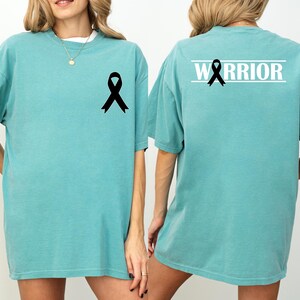 May include: Seafoam green t-shirt with a black ribbon graphic on the front and back. The back of the shirt features the word "WARRIOR" in white block letters above the ribbon. The shirt is a comfortable, oversized fit.