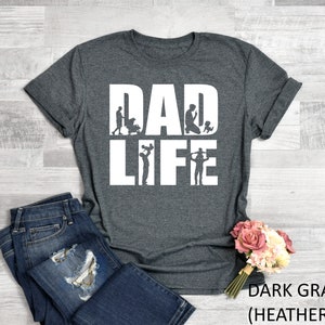 Peut inclure: T-shirt gris fonc&eacute; chin&eacute; avec le texte "DAD LIFE" en blanc. Le texte est entour&eacute; de silhouettes d'un p&egrave;re &agrave; diff&eacute;rents stades de la paternit&eacute;, de la tenue d'un b&eacute;b&eacute; &agrave; la  jouer avec un enfant.