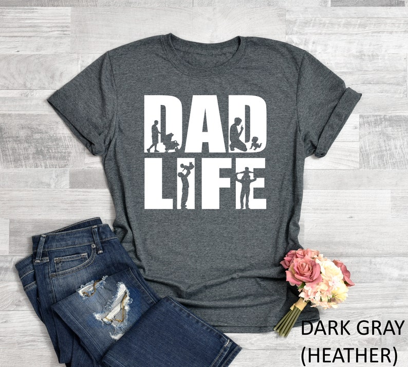 Peut inclure: T-shirt gris fonc&eacute; chin&eacute; avec le texte "DAD LIFE" en blanc. Le texte est entour&eacute; de silhouettes d'un p&egrave;re &agrave; diff&eacute;rents stades de la paternit&eacute;, de la tenue d'un b&eacute;b&eacute; &agrave; la  jouer avec un enfant.
