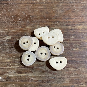 May include: A group of eight white antler buttons with two holes each. The buttons are arranged in a pile on a wooden surface.