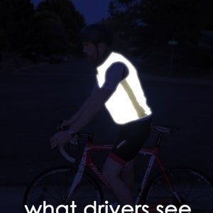 May include: A cyclist wearing a reflective vest rides a red and white road bike at night. The vest is bright yellow and highly visible. The text "what drivers see" is at the bottom of the image.