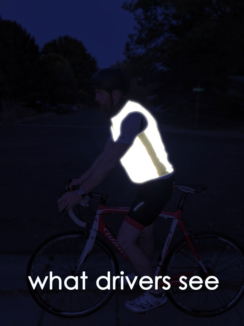 May include: A cyclist wearing a reflective vest rides a red and white road bike at night. The vest is bright yellow and highly visible. The text "what drivers see" is at the bottom of the image.