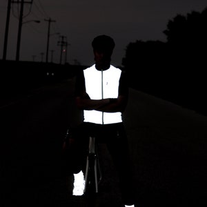 May include: A person wearing a reflective vest, pants, and shoes, standing on a dark road. The vest is white and reflective, with a zipper down the front. The pants and shoes also have reflective elements, enhancing visibility in low-light conditions.