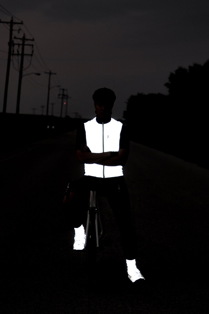 May include: A person wearing a reflective vest, pants, and shoes, standing on a dark road. The vest is white and reflective, with a zipper down the front. The pants and shoes also have reflective elements, enhancing visibility in low-light conditions.