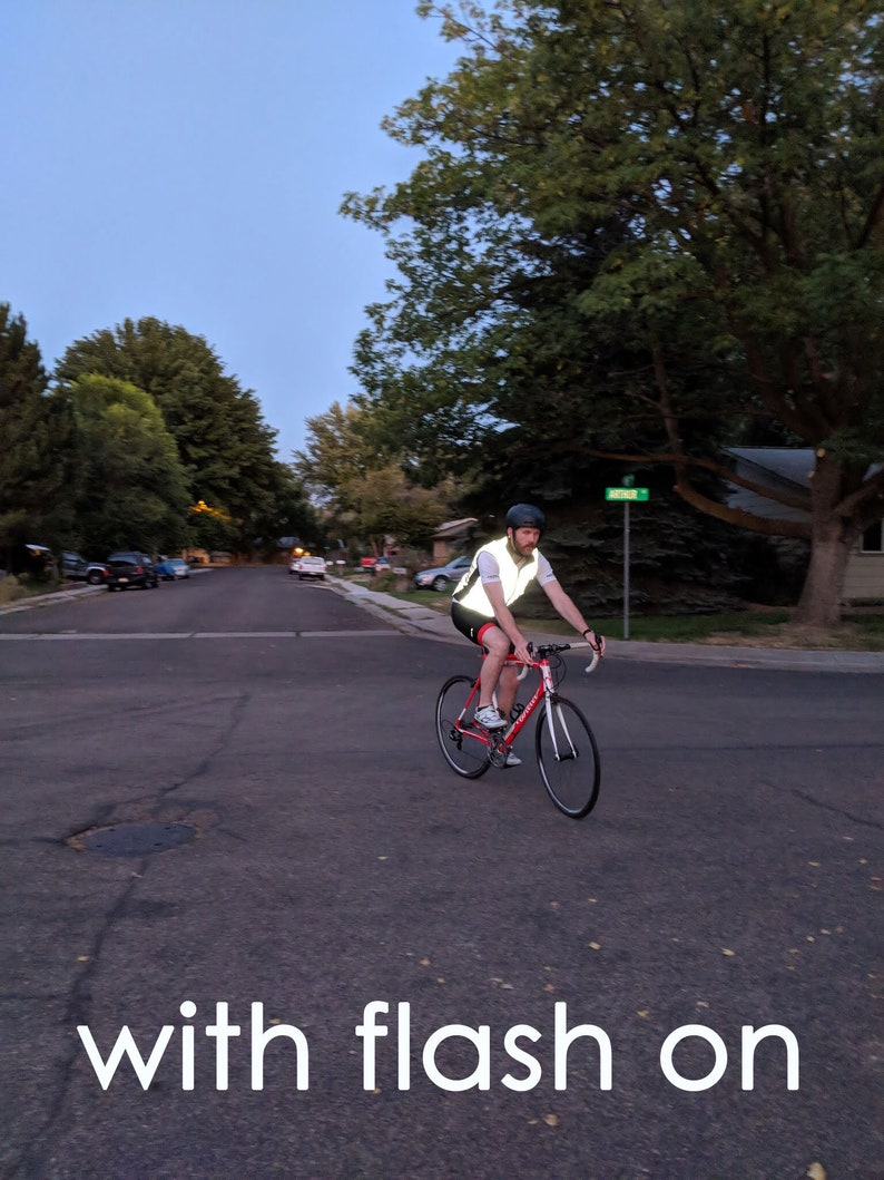 May include: A person riding a red road bicycle on a paved street at dusk. The cyclist wears a reflective vest and helmet for safety. The image includes the text "with flash on".